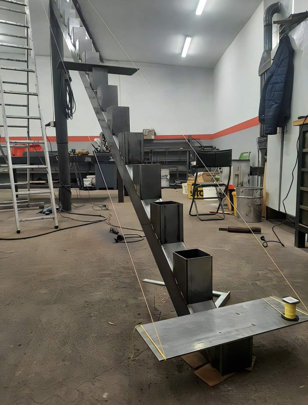 Metal staircase under construction in a workshop; unfinished steps, angled beam, support cables.