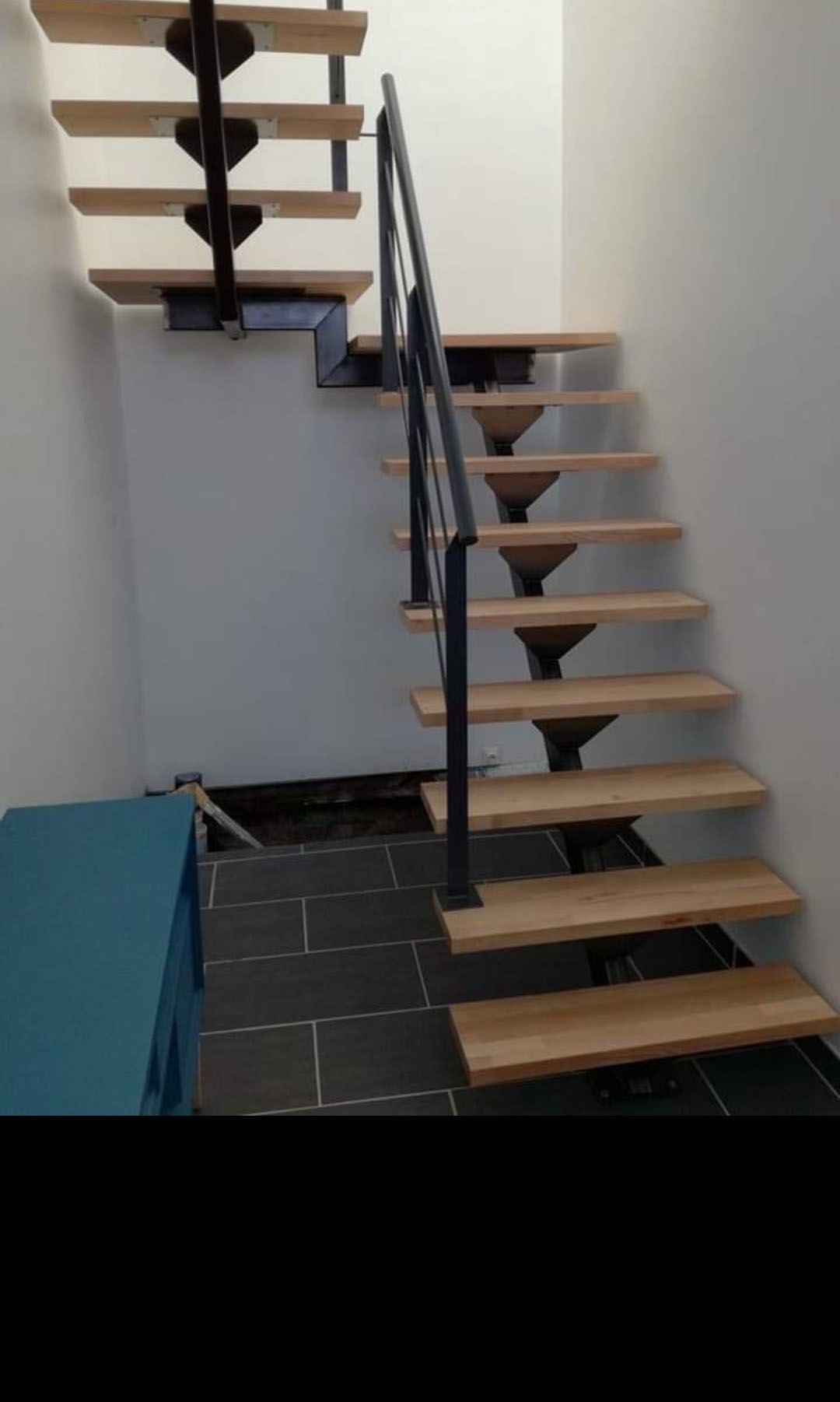 Wooden staircase with black metal supports and railing against white walls.