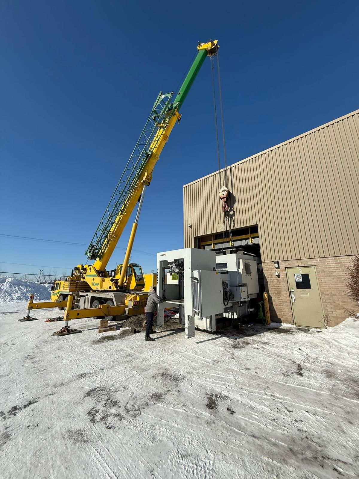 A yellow crane lifts machinery into a warehouse. Snowy ground, blue sky.