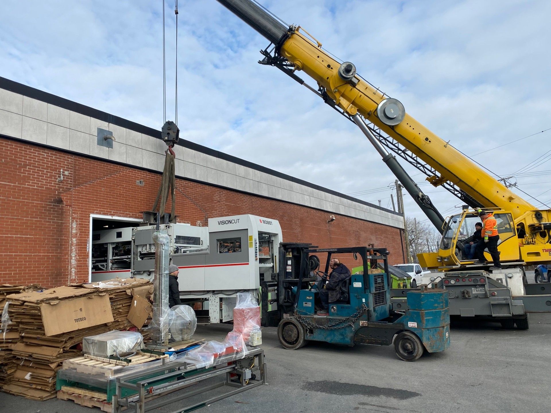 A yellow crane lifts equipment from a truck, workers nearby.