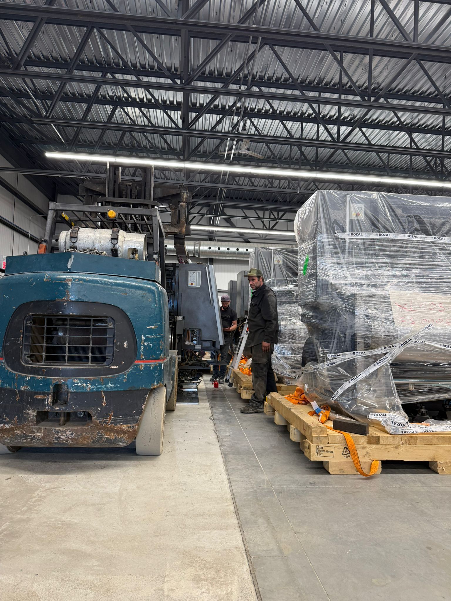 Forklift loading large packaged items in a warehouse. Two workers supervise the operation.