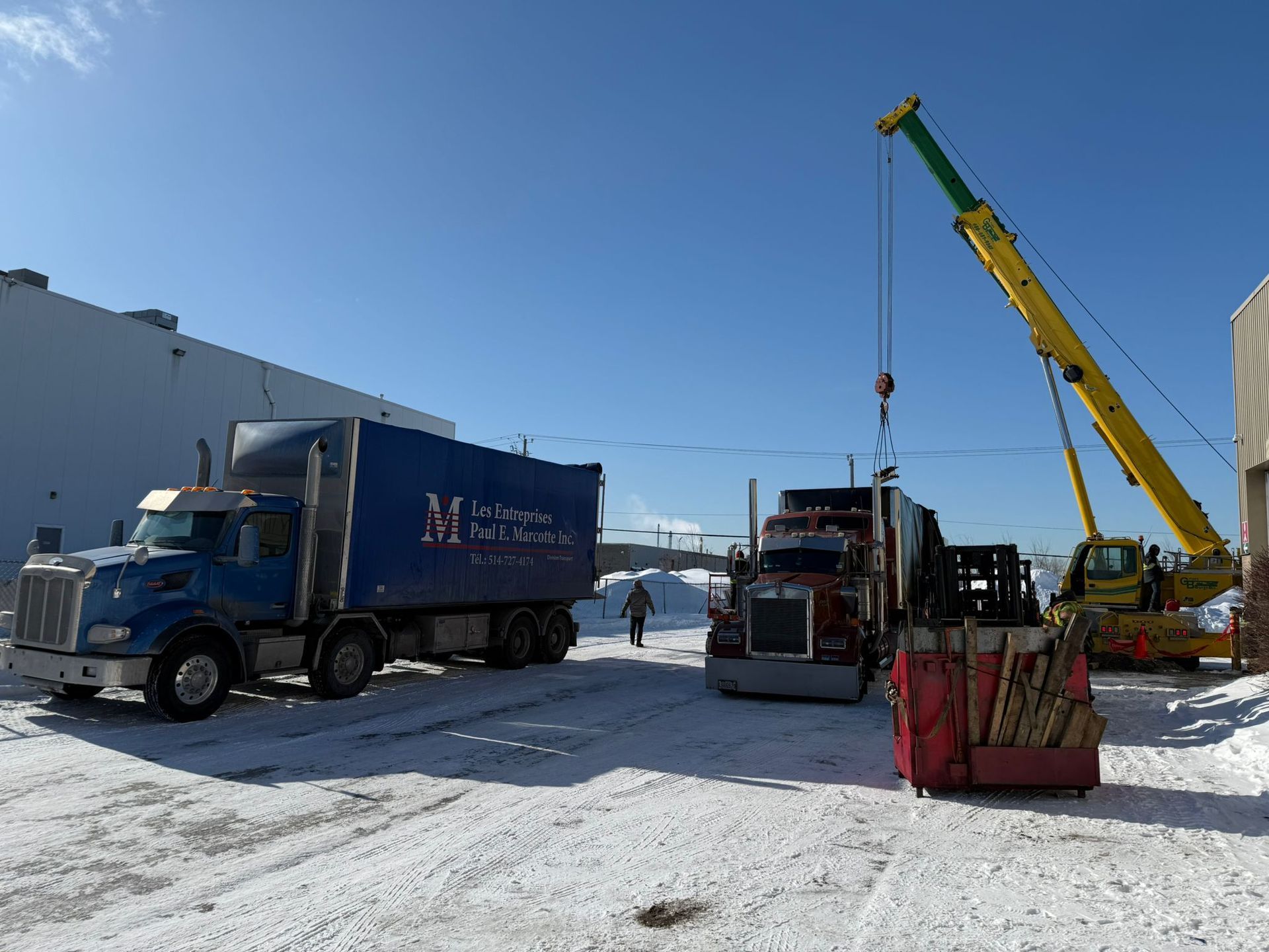 Trucks being loaded by a crane in a snowy parking lot; blue sky.