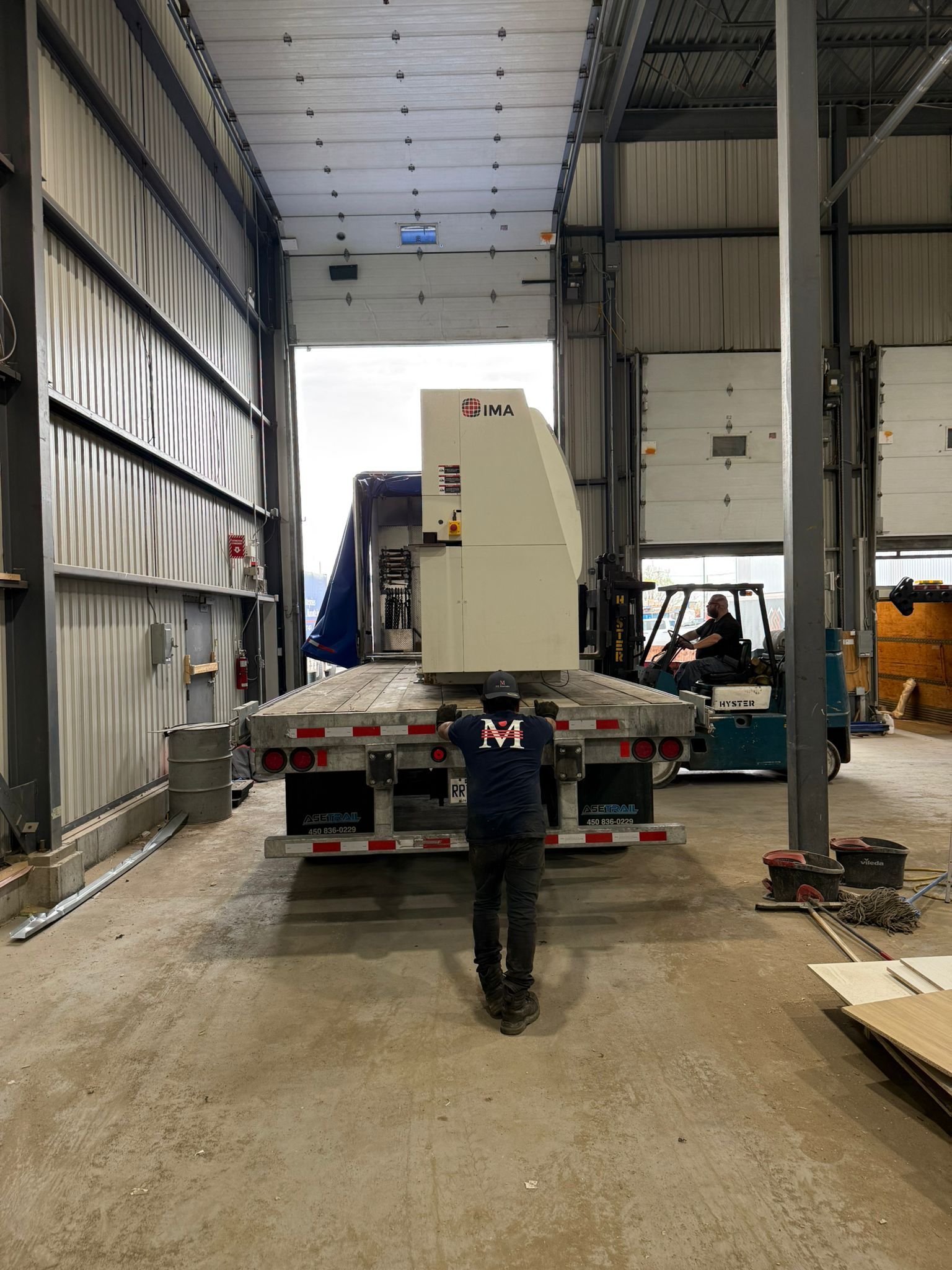 A worker guides machinery off a truck. A forklift waits. Inside a warehouse.