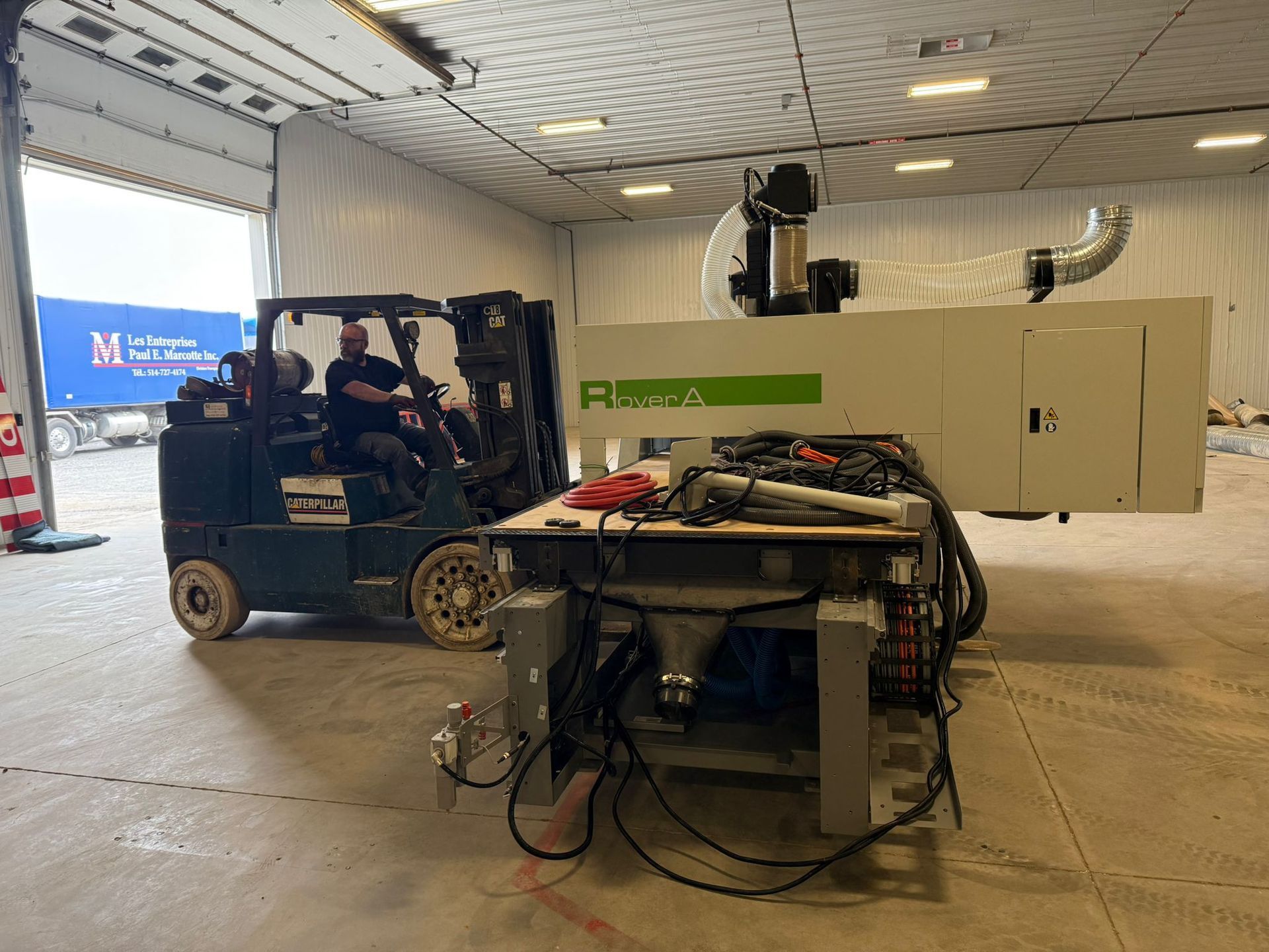 Forklift moving machinery in warehouse, with a person driving. A truck is visible outside.
