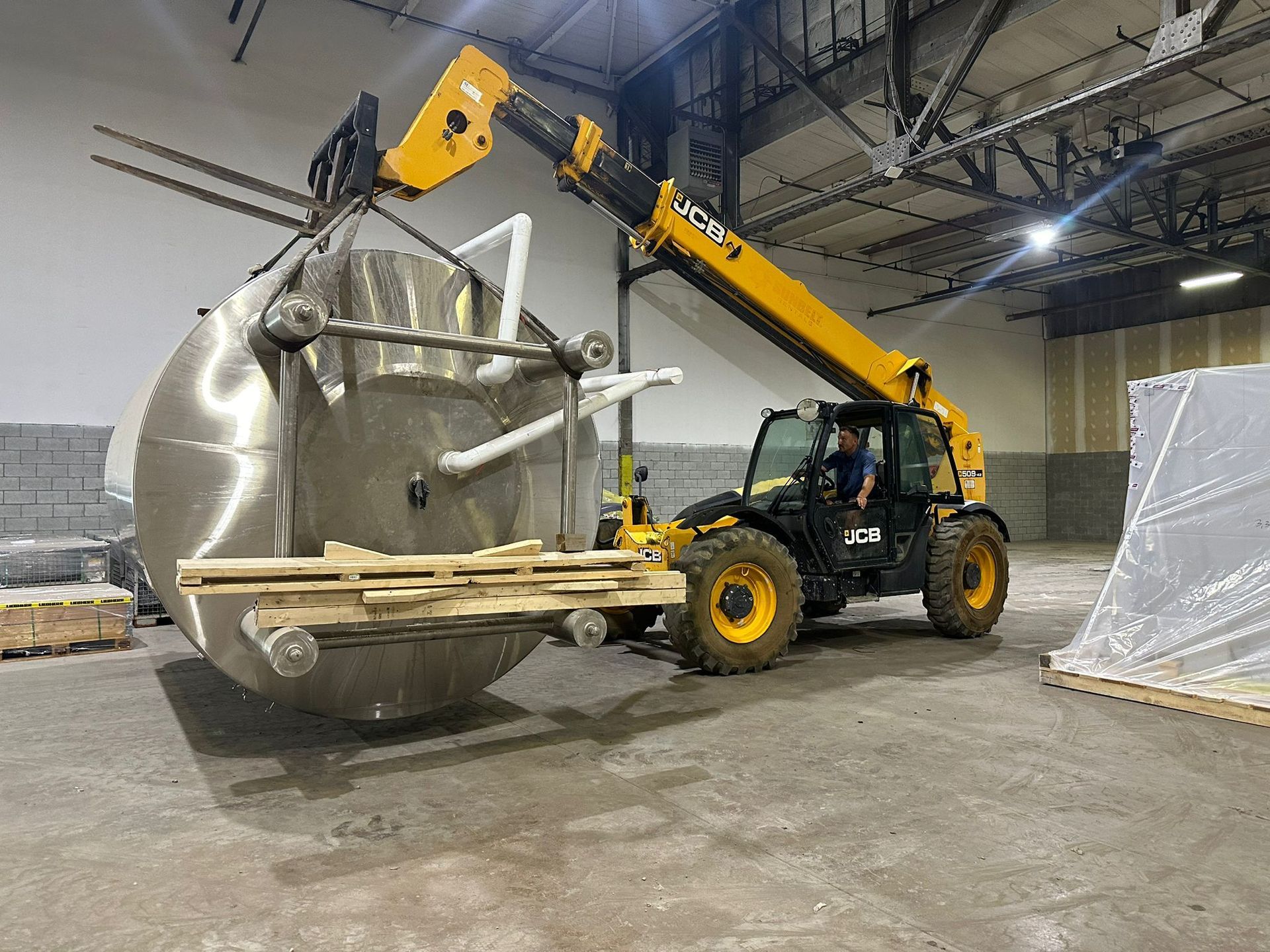 A telehandler lifts a large, silver industrial tank on a wooden pallet inside a warehouse.