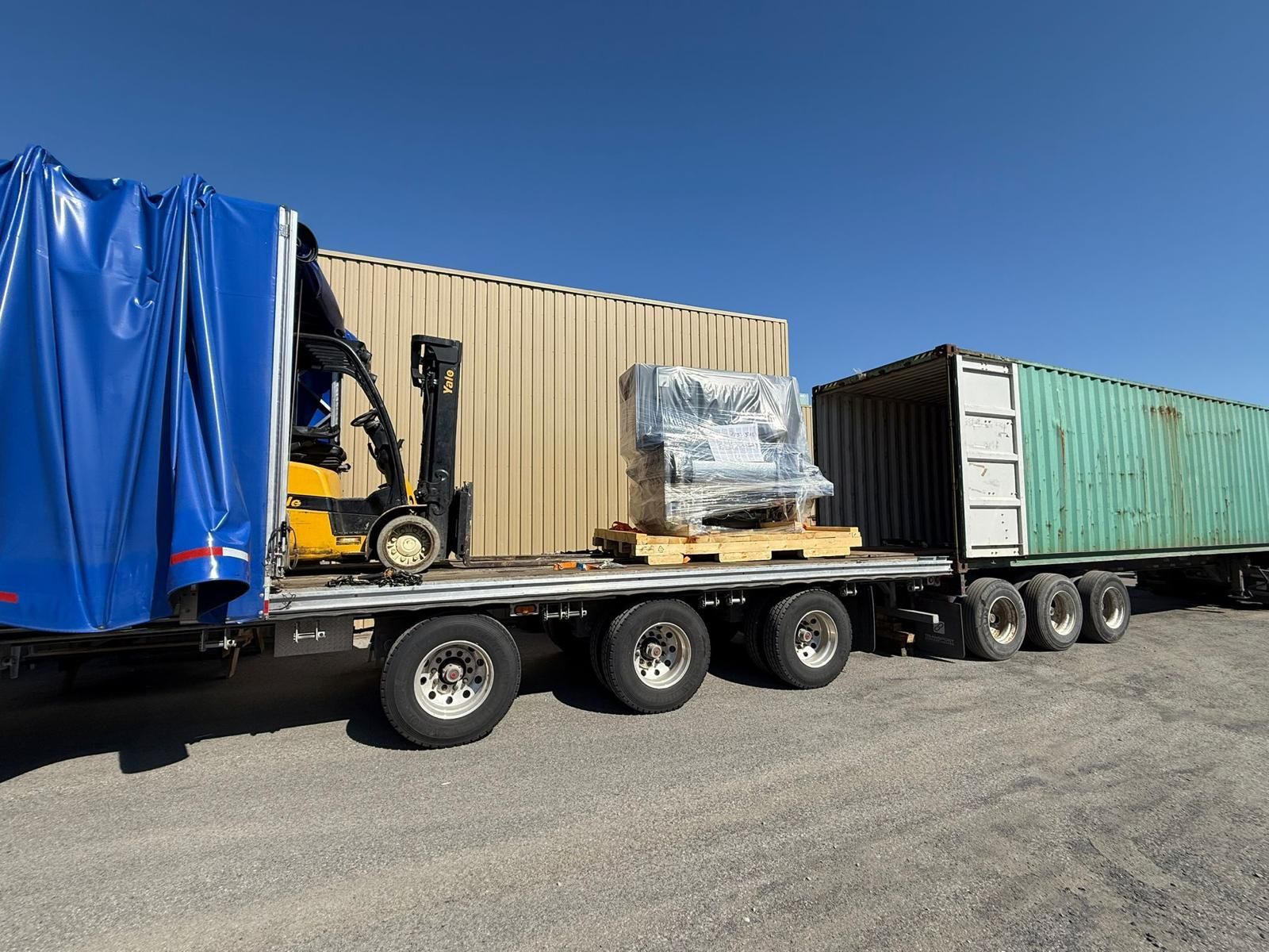 Forklift loading crate onto a flatbed truck next to a shipping container under a blue sky.
