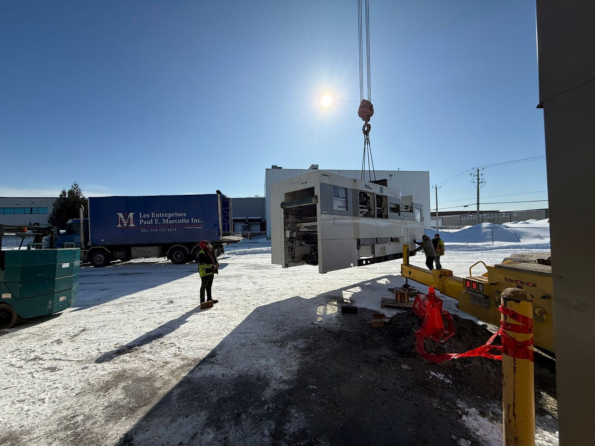 A crane lifts a large industrial unit outside on a snowy day; a truck and workers are nearby.