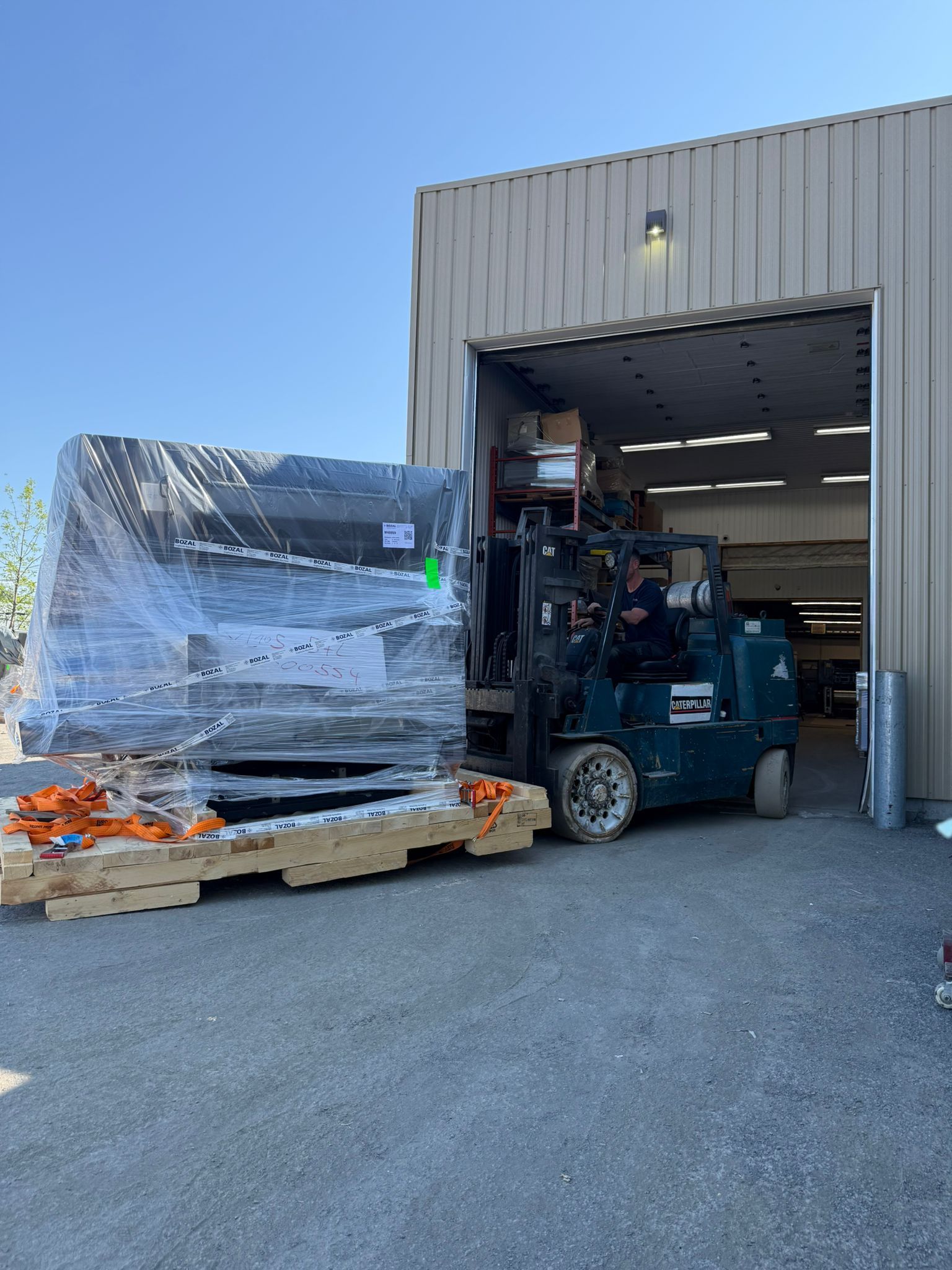 Forklift loading a large, wrapped pallet into a warehouse. The forklift is green. Sunny day.