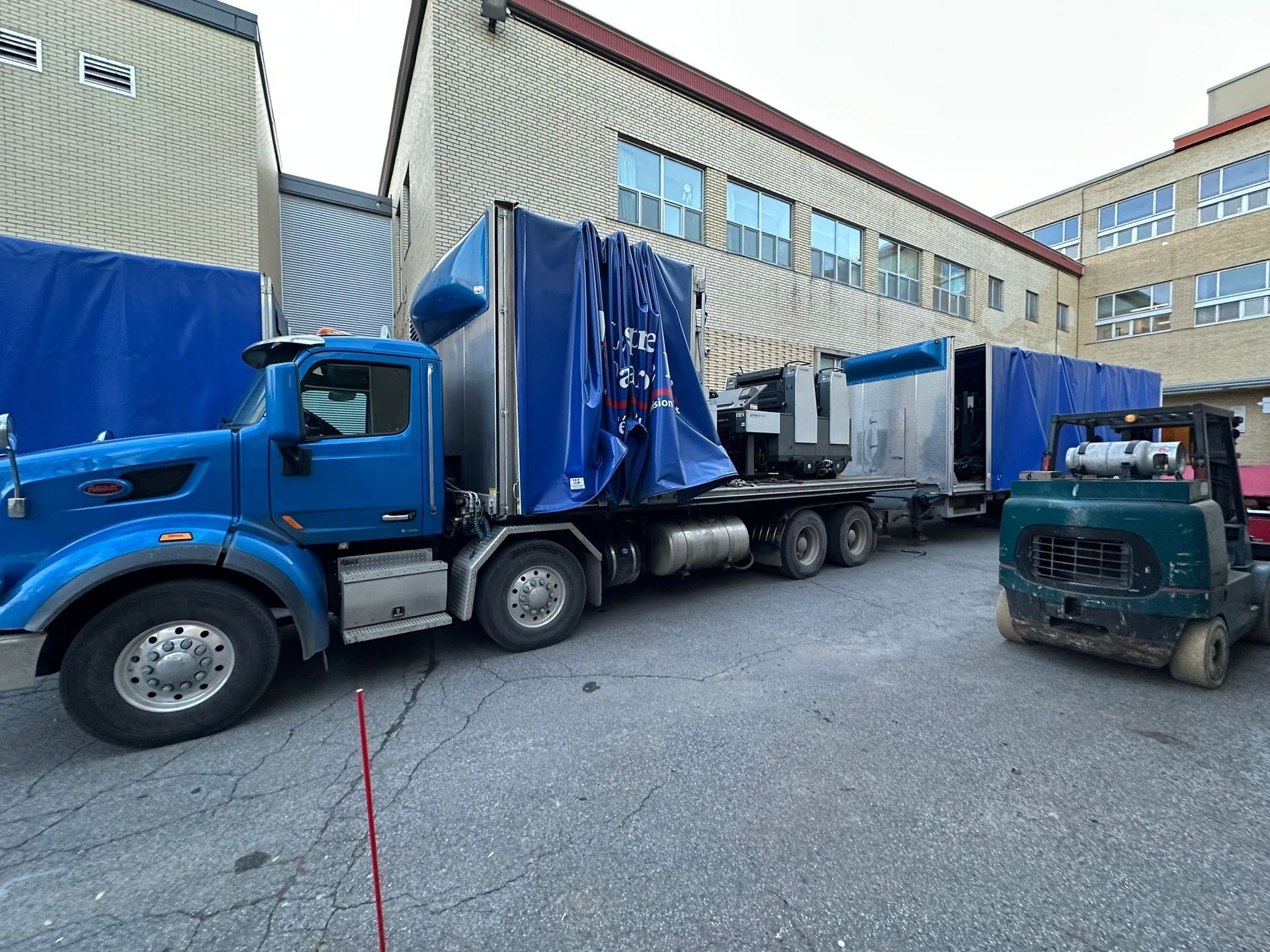 Blue semi-truck parked near a building, loading/unloading with a forklift.