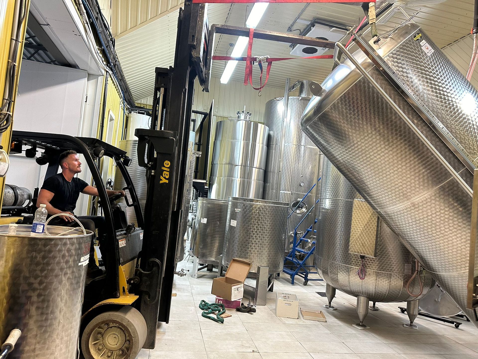 Man operating forklift lifting large metal tank in a warehouse with other tanks.