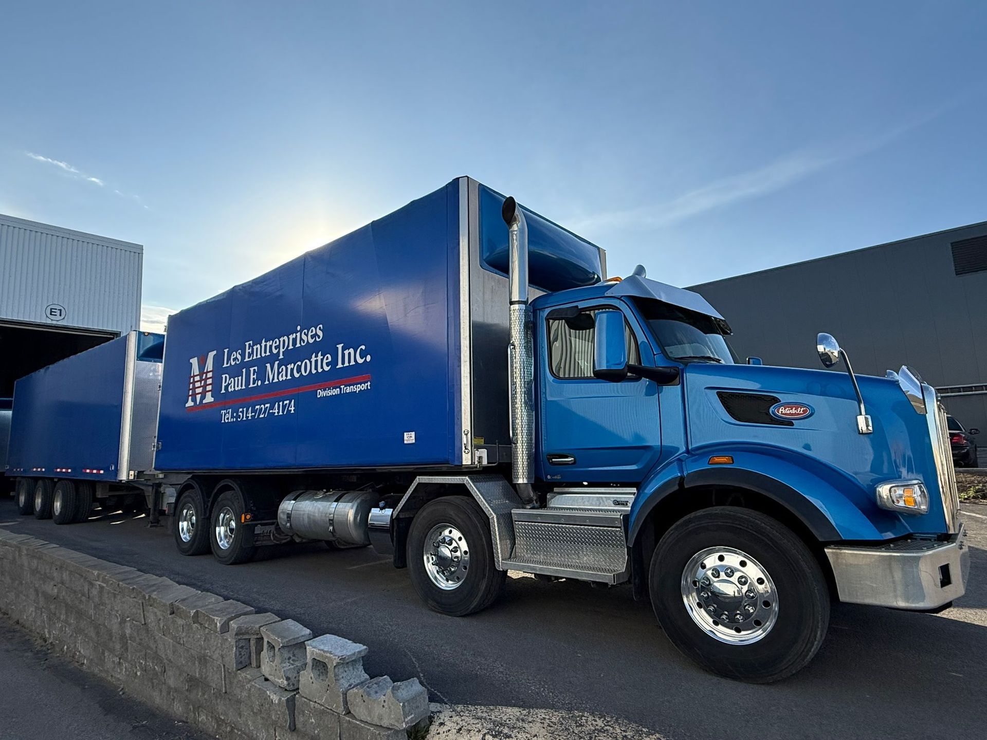 Blue semi-truck with trailer, parked at a loading dock, logo on side.