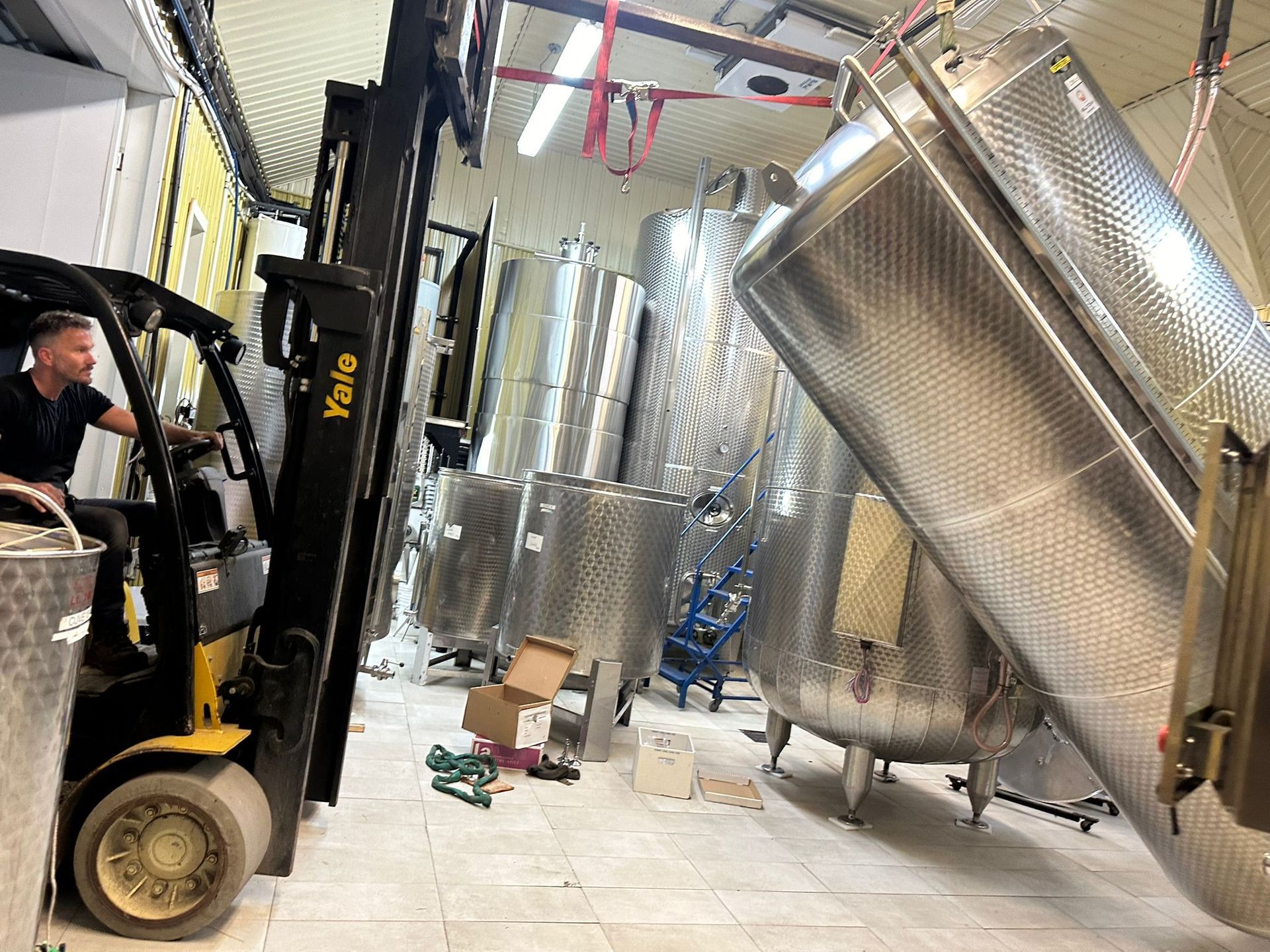 Man operating a forklift, tilting a large steel tank inside a warehouse with several other tanks.