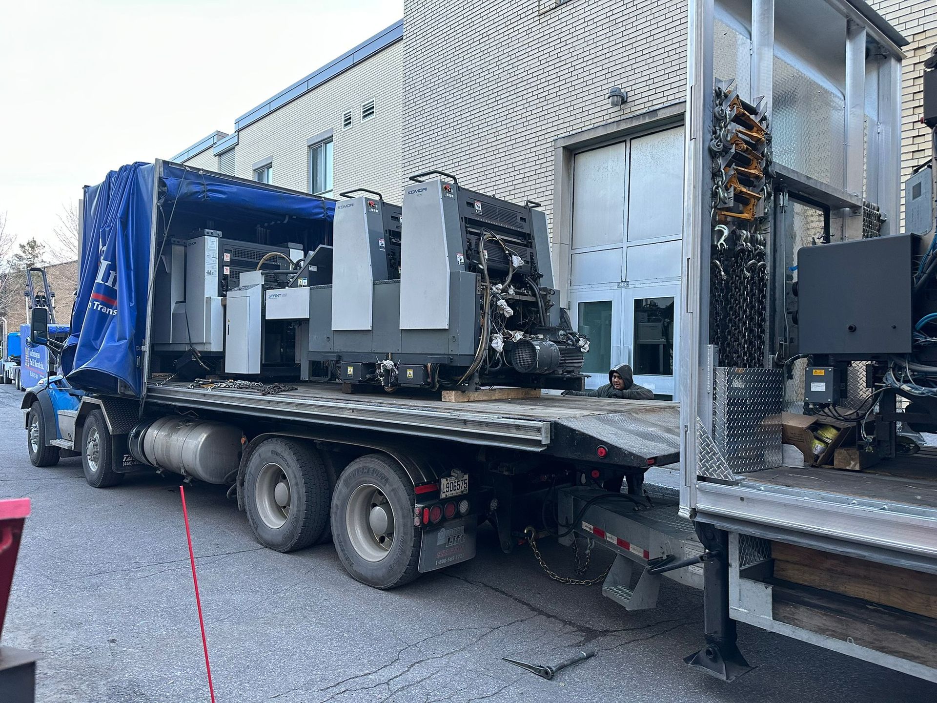 A large printing press on a flatbed truck being unloaded near a building with a loading dock.