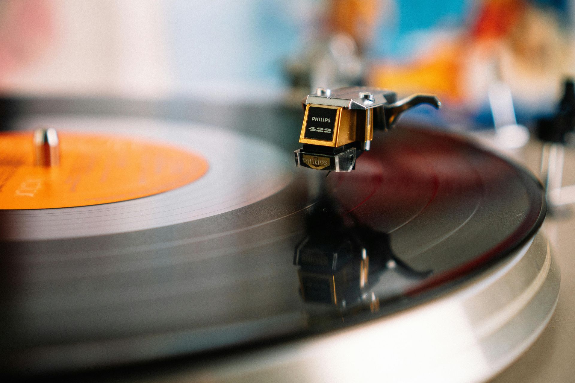 Turntable playing vinyl record, close-up. Black record with orange label. Tone arm near center.