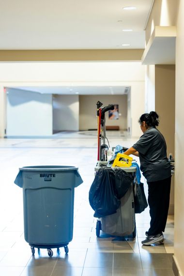 Custodian cleaning a hallway, pushing a cart with supplies beside a blue trash bin