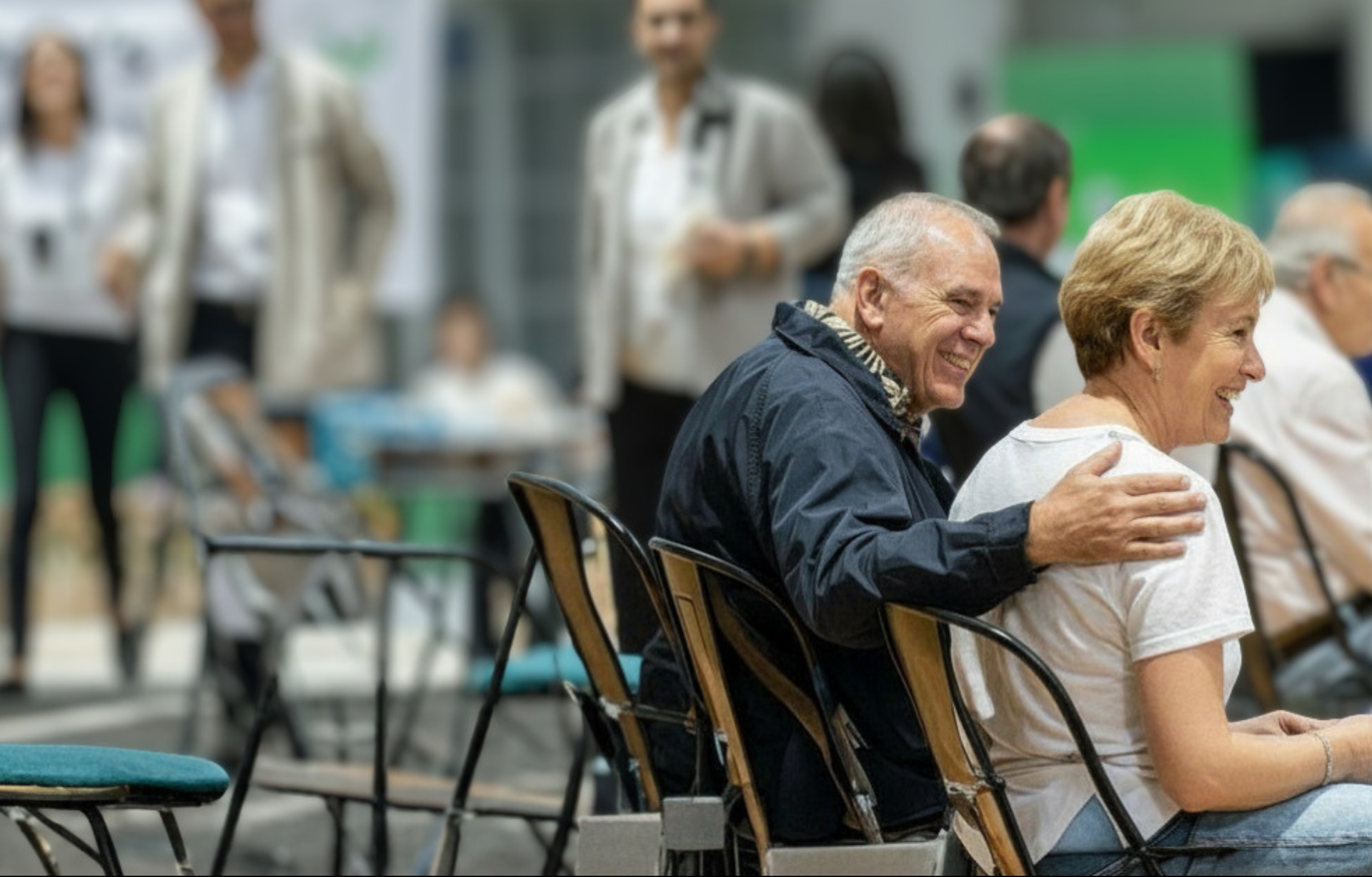 Older man smiling with arm around seated woman in white shirt, in a conference hall with people blurred in background