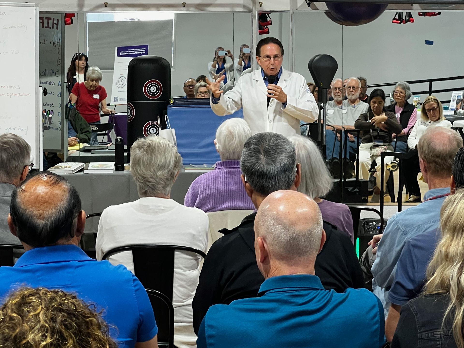 Speaker addressing seated audience in a hall, with boxing equipment and posters in the background