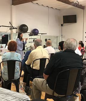 Audience seated indoors watching a presentation beside exercise equipment and wall-mounted screens.