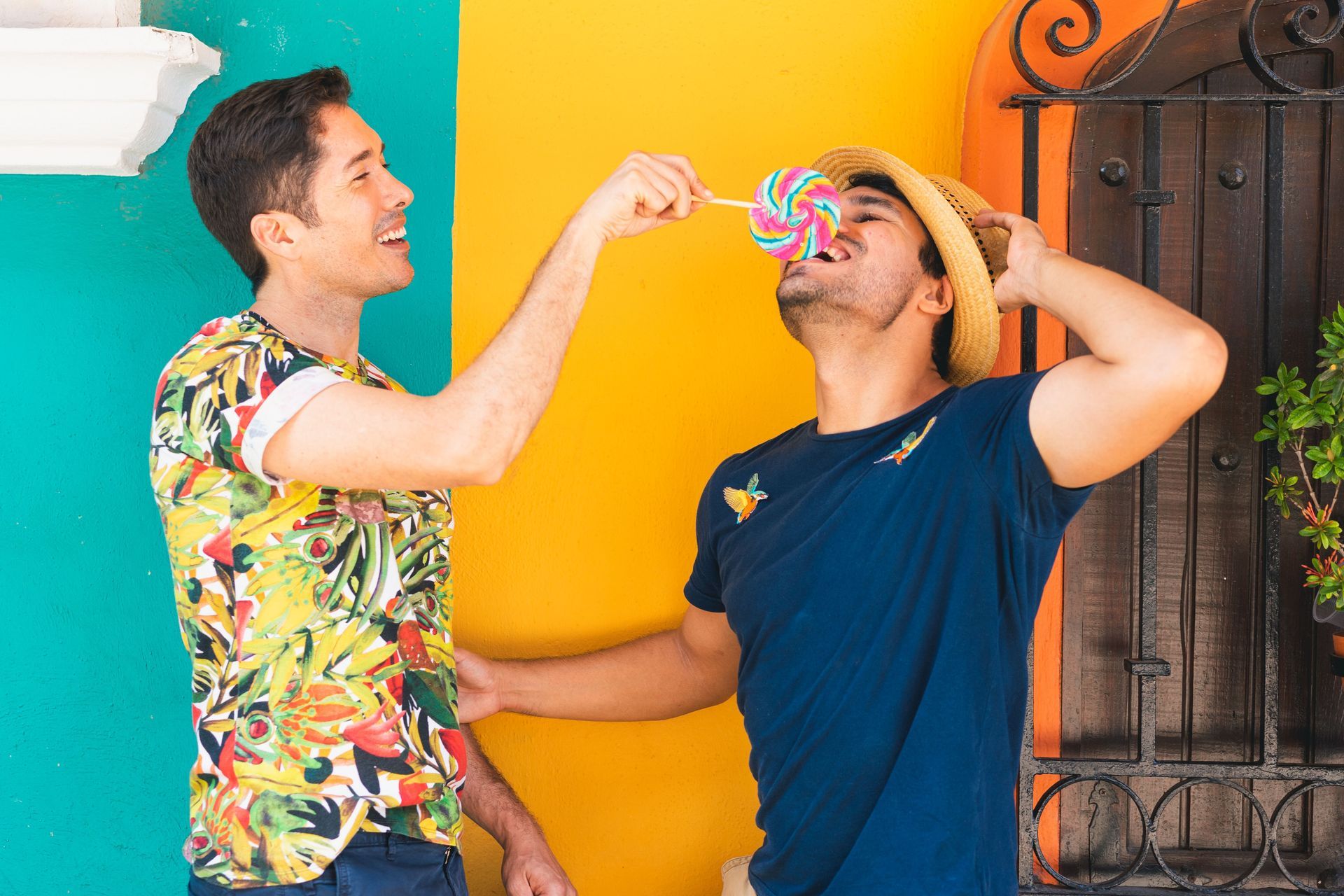 Two men sharing a colorful drink at a bright yellow wall, smiling and laughing together.