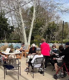 People seated outdoors in a park-like setting, gathered around tables and chairs on a sunny day