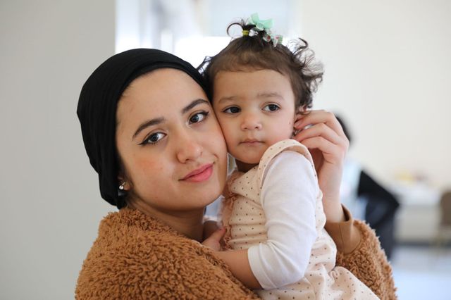 Woman in black hat holds a young child in a white shirt and dress, both looking at the camera.