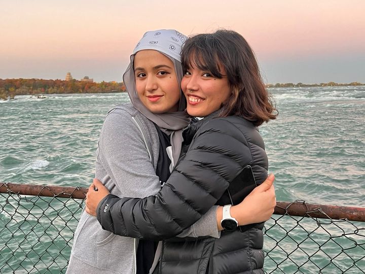 Two women embrace near a body of water; one wears a hijab, the other a black jacket.
