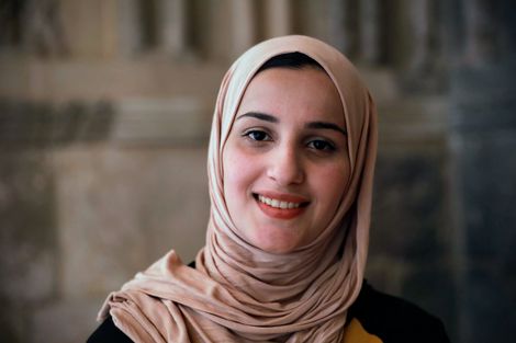 Woman in a beige hijab smiles in front of a stone wall.