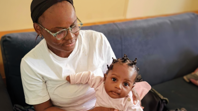 Woman holding baby on a blue couch. The woman wears glasses and a white shirt, looking at the baby.
