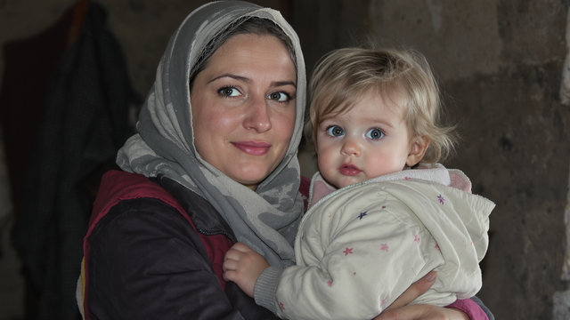 Woman in headscarf holding a baby; both looking at the camera indoors.