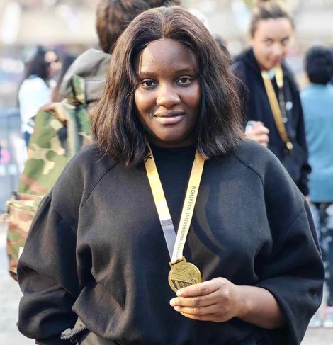 Woman with a gold medal smiling at the camera, wearing black clothing, outdoors.