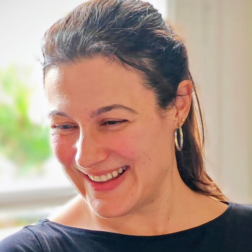 Woman smiling broadly, looking down. Wearing hoop earrings, dark top, light background.