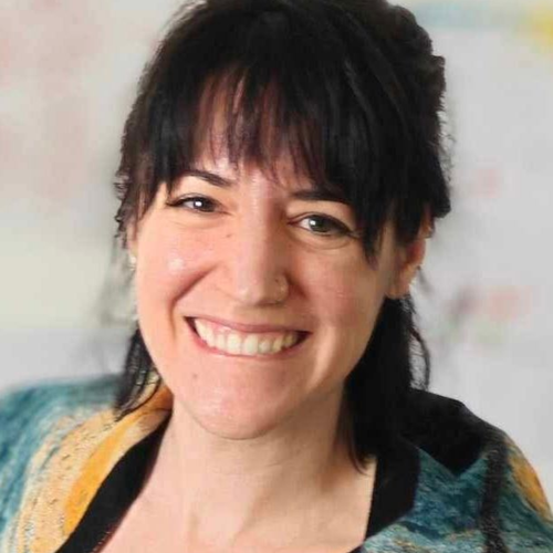 Woman with dark hair, smiling, wearing a colorful patterned jacket, looking directly at the camera.