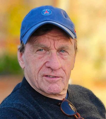 Man wearing a blue cap with a logo, looking at the camera. He wears a dark sweater.