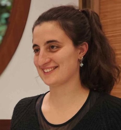 Woman with dark hair in a ponytail smiles, wearing a black sweater and small earrings.