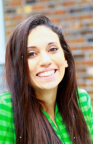 Woman with long brown hair smiles widely in front of a brick building, wearing a green plaid shirt.