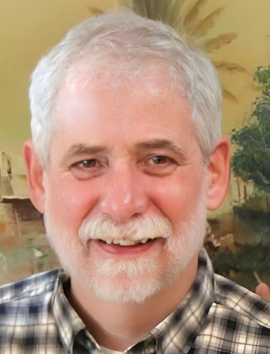 Bearded man with gray hair smiles, wearing a plaid shirt; soft focus background.