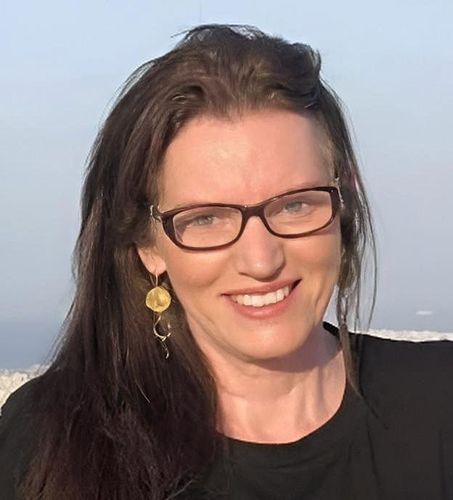 Woman with long brown hair wearing glasses smiles. She wears a black top and gold earrings. Background is blurred.