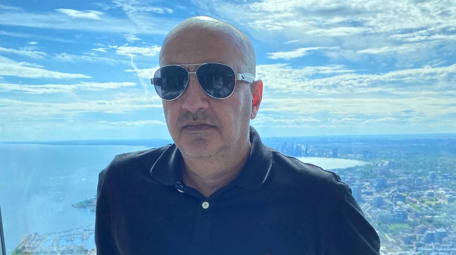 Man wearing sunglasses and a black polo shirt, standing in front of a window with a city view.