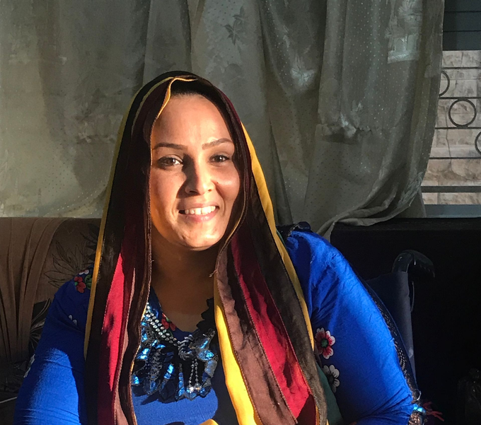 Woman in blue embroidered dress and colorful headscarf smiles.