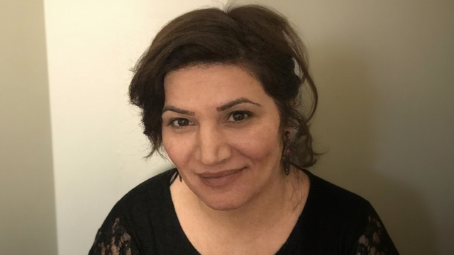 Woman with dark hair smiles, wearing a black top, against a neutral wall.