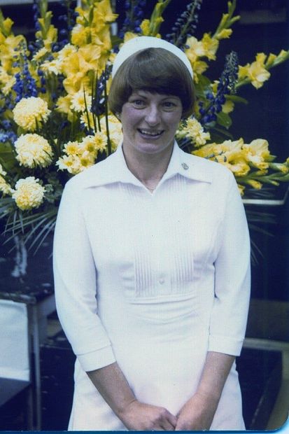 Woman in a white uniform and cap smiles in front of yellow and blue flowers.