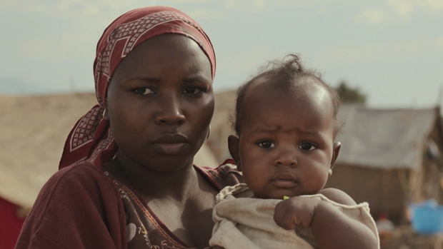 Woman in a headscarf holds a baby. Both look somber, outside with tents in the background.