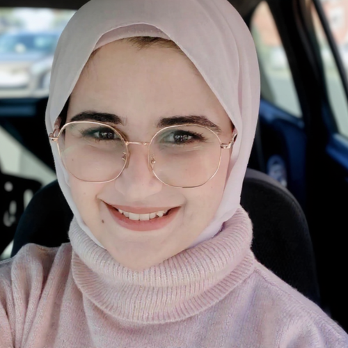 Woman wearing a pink hijab, glasses, and turtleneck smiles in a car.