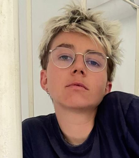 Person with short, spiky blonde hair, wearing glasses, and a dark blue shirt, leaning towards the camera.