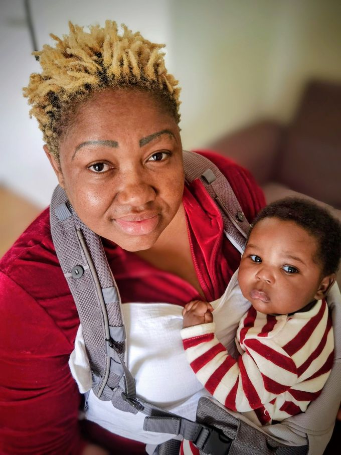 Woman with blonde-tipped hair holds a baby in a carrier; both gaze at the viewer.