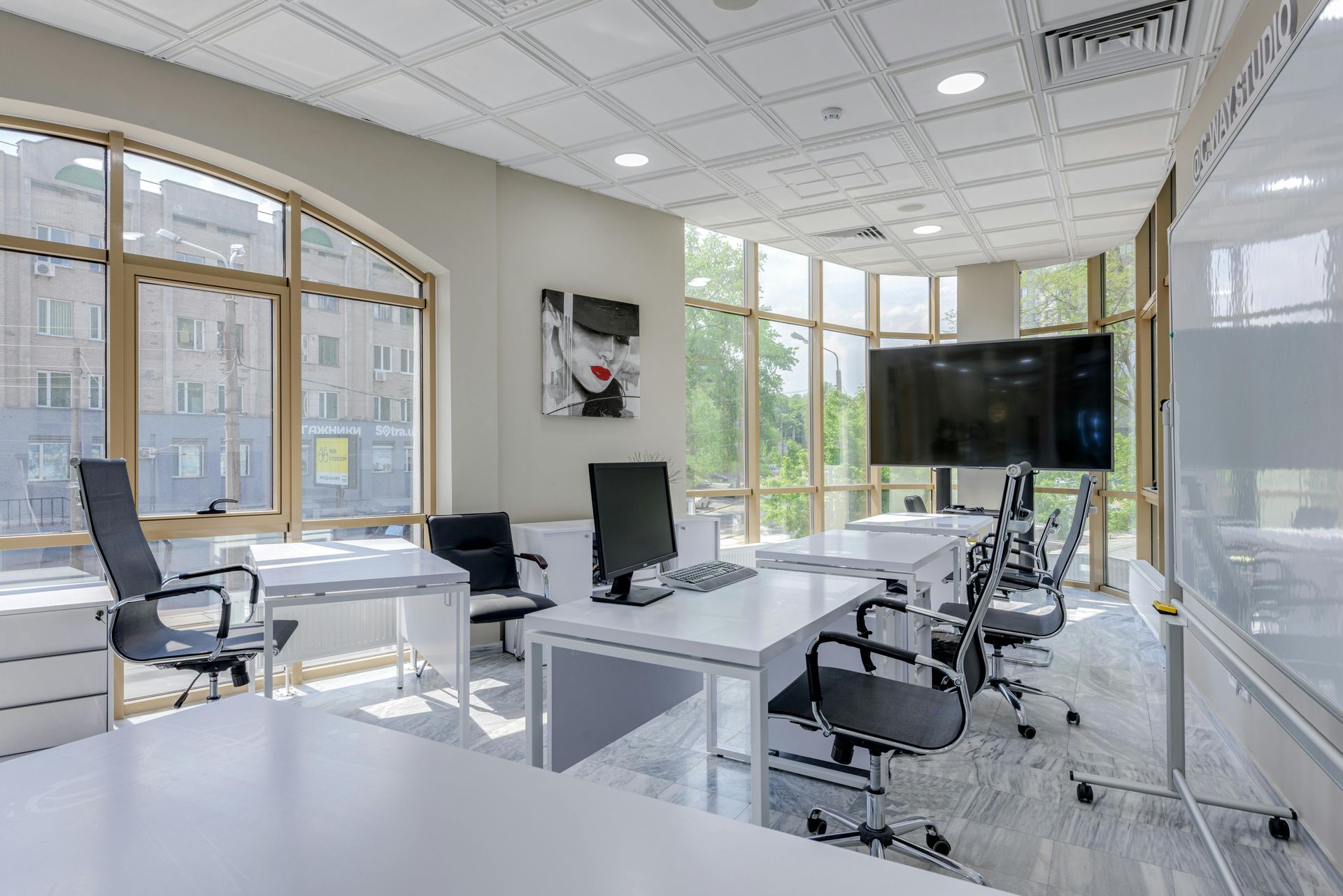 Modern office with white desks, chairs, large windows, and a large TV. Artwork on wall.