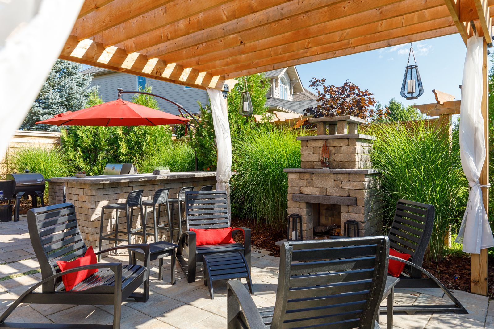 A patio with chairs , umbrellas and a fireplace under a pergola.