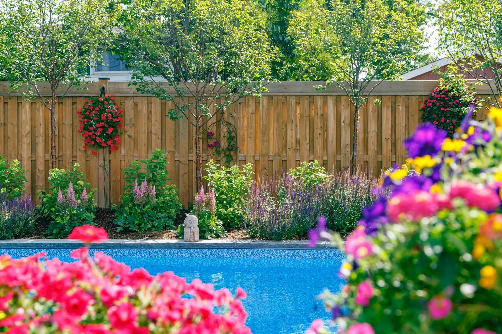 A wooden fence surrounds a swimming pool surrounded by flowers and trees.