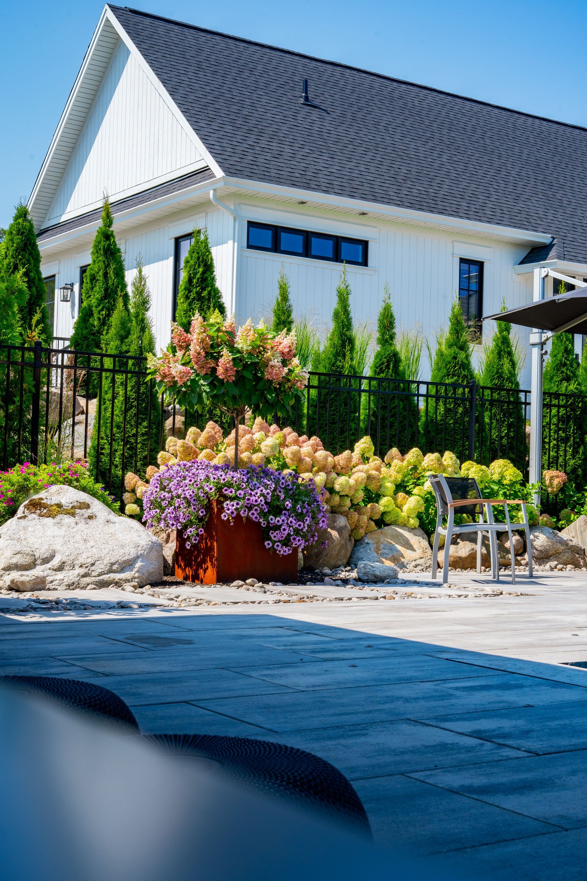 A white house with a black roof is surrounded by trees and flowers.