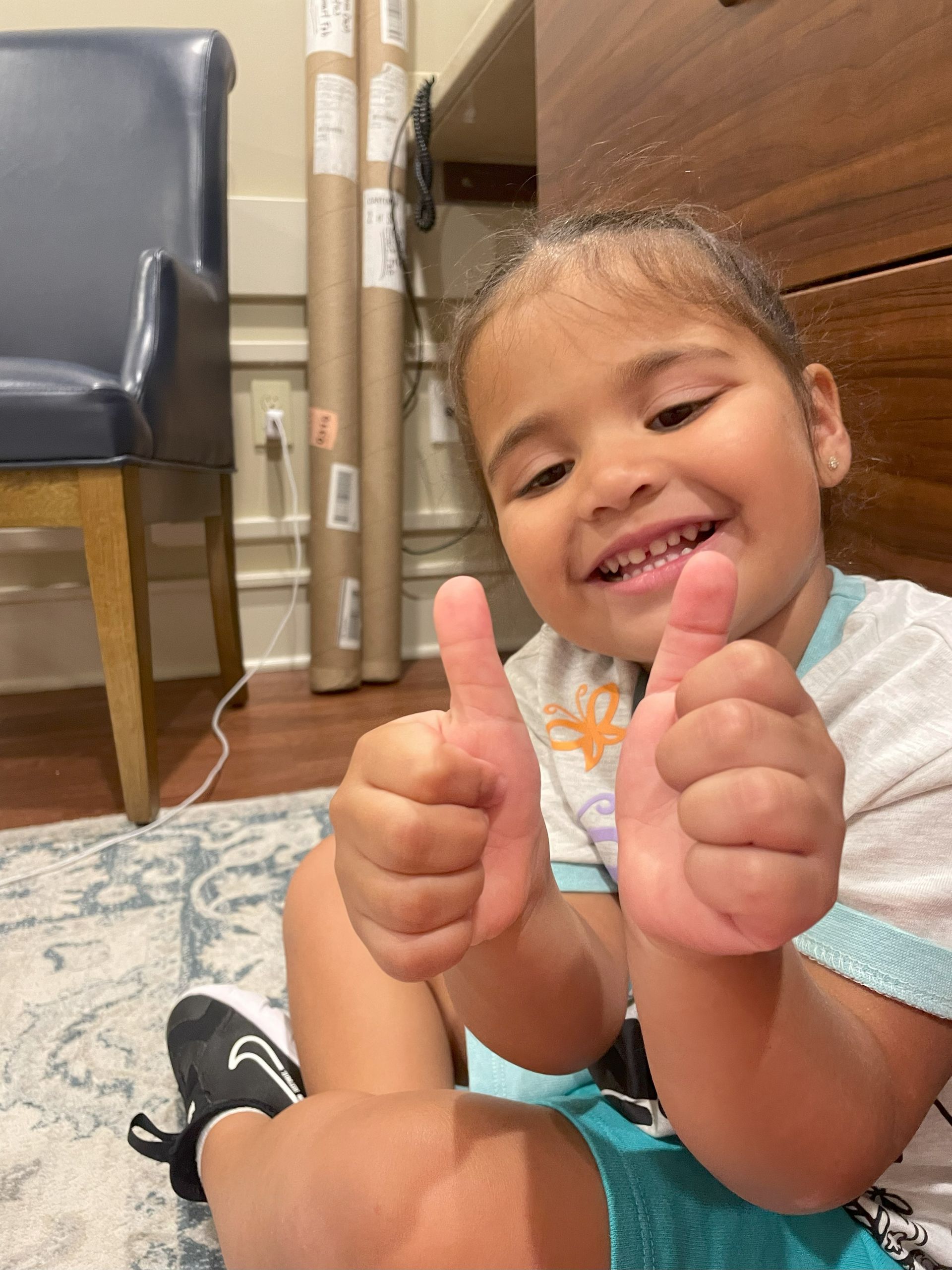 A little girl is sitting on the floor giving two thumbs up.